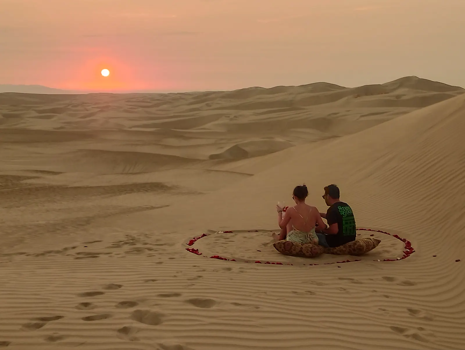 Pareja brindando dentro de un círculo de pétalos en las dunas, al atardecer
