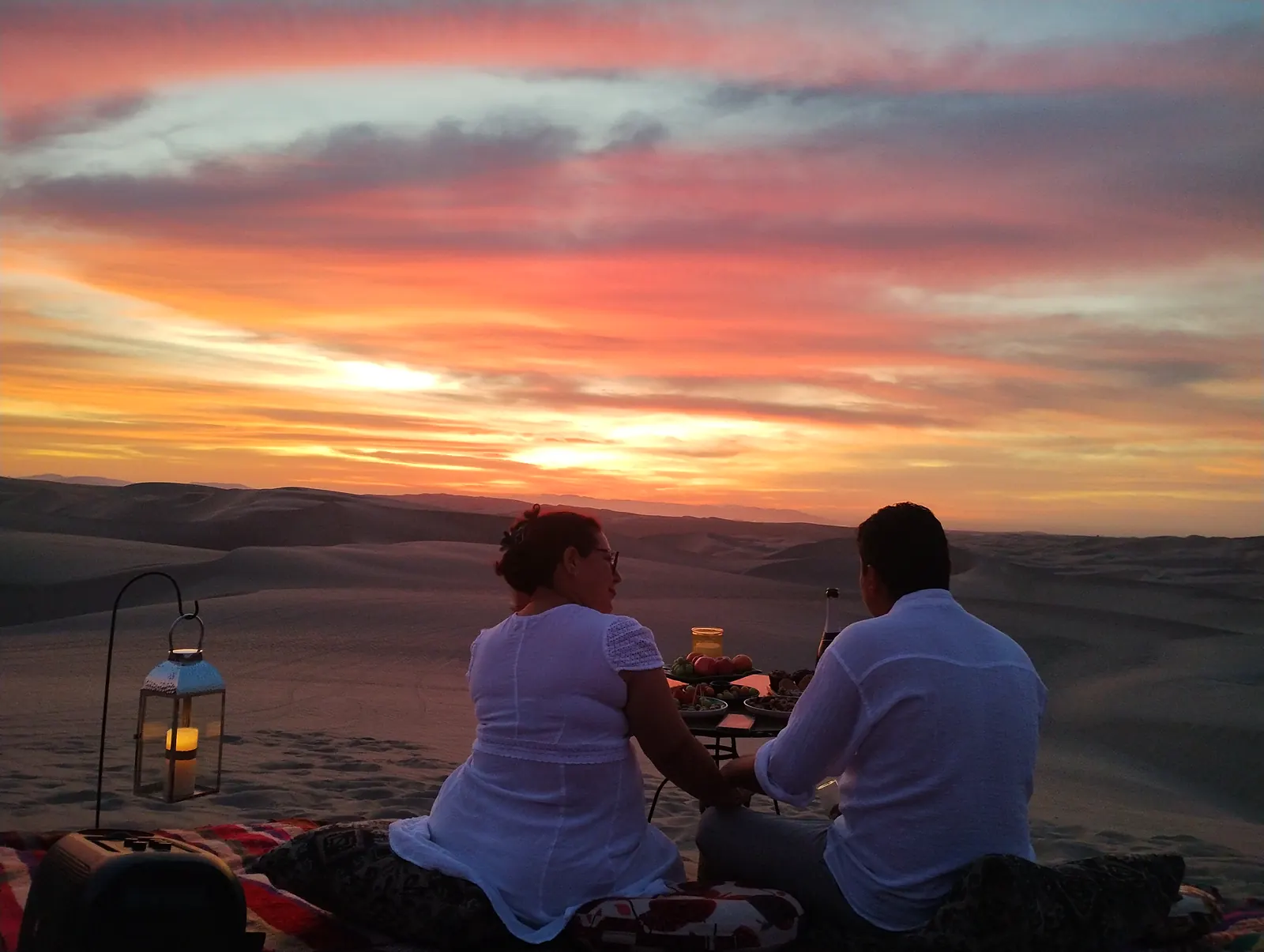 Pareja frente al cielo encendido del atardecer, con un farol en las dunas